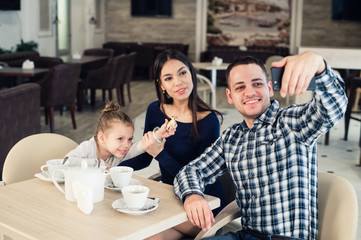 family, parenthood, technology people concept - happy mother, father and little girl having dinner taking selfie by smartphone at restaurant