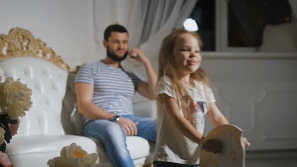 Close-up of father and daughter playing together. Happy little girl is riding a rocking horse and laughing with father smiling and sitting on the big white sofa. - Powered by Adobe