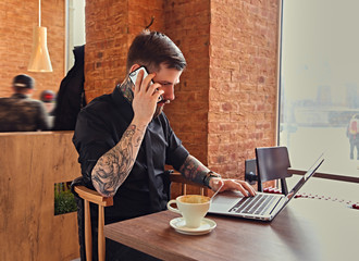 A man talking by smartphone and using a laptop in a coffee shop.