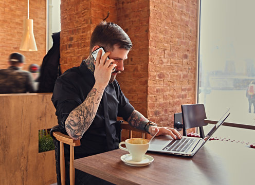 A man talking by smartphone and using a laptop in a coffee shop.