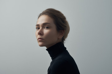 Dramatic portrait of a young beautiful girl with freckles in a black turtleneck on white background in studio - 129438550