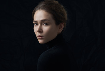 Dramatic portrait of a young beautiful girl with freckles in a black turtleneck on black background...