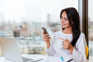 Cheerful young businesswoman with laptop sitting and using mobile phone