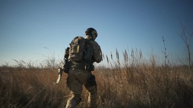 Soldier in full NATO ammunition walks in field, squats down, aims with a rifle