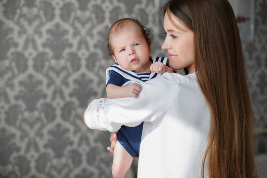 Portrait of mother and child,beautiful mom,brunette with long straight hair,dressed in a white shirt,is holding his newborn son,the portrait is painted in the room with the grey Wallpaper