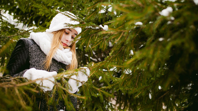 young woman in a forest with fir and pine trees on the street, hands touch the branches of trees, considering them. peridium winter, warm clothes, knitted mittens and scarf white