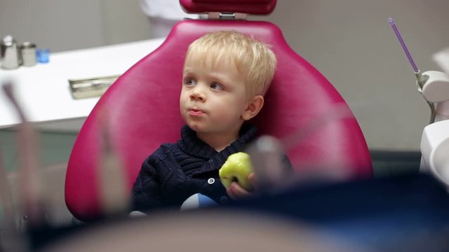 Small child sitting in the dental chair and eats a green apple. Examination of the teeth at the dentist.