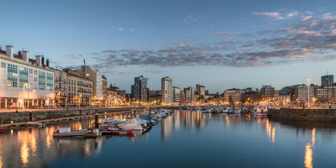 Fototapeta na wymiar Yatchs and pier in leisure port on maritime fishing district of Gijon, Spain, Europe. Beautiful reflection on calm sea water of boats, buildings, sky at dusk at touristic cultural travel destination.
