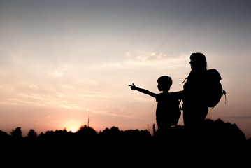 Fototapeta na wymiar Silhouettes of mother and child hiking at sunset. Boy pointing at something distant. Summer vacation in mountains. Traveling with little kids. Mom and son with backpacks standing on the hill edge.
