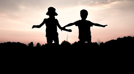 Silhouettes of children jumping off a cliff at sunset. Little boy and girl jump raising hands up high. Brother and sister having fun in summer. Friendship, freedom concept.