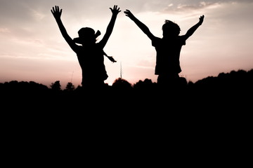Silhouettes of kids jumping off a cliff at sunset. Little boy and girl jump raising hands up high. Brother and sister having fun in summer. Friendship, freedom concept