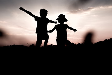 Silhouettes of children jumping off a hill at sunset. Little boy and girl jump raising hands up high. Brother and sister having fun in summer. Friendship, freedom concept