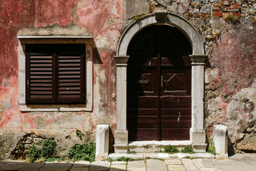 Fototapeta na wymiar Shabby wall of the building with a maroon door and window with shutters in Porec, Croatia.