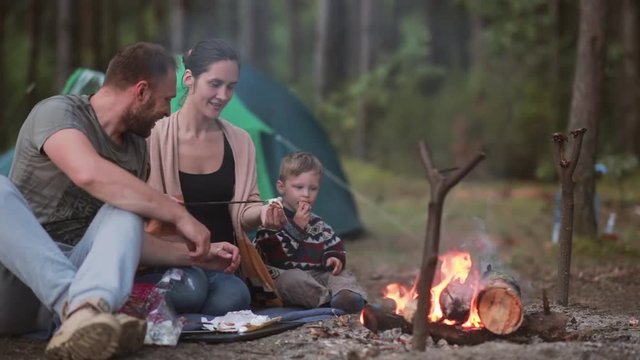 Family rest in the nature. Husband and wife with their little son are cooking marshmallows on open fire and eating them.
