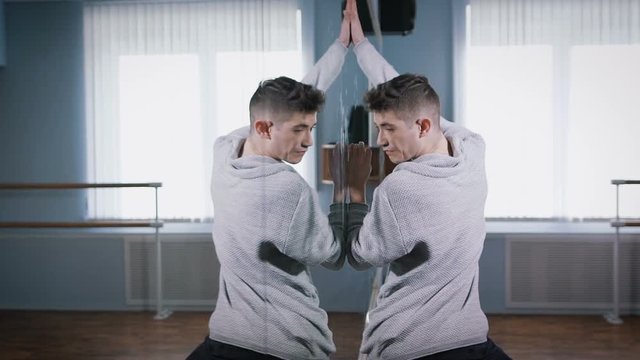 Close up of handsome dancer in grey pullover and black trousers dancing near the mirror on the wall. Young man is performing modern dance with passion near the mirror.