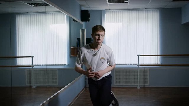 Close up of young hip-hop dancer in black trousers and white shirt performing urban dance standing near the mirror. B-boy is jumping and moving his arms and legs showing freestyle and modern dance.