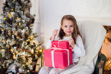 Happy girl opening Christmas gifts by a decorated fireplace in cozy light living room on Xmas eve