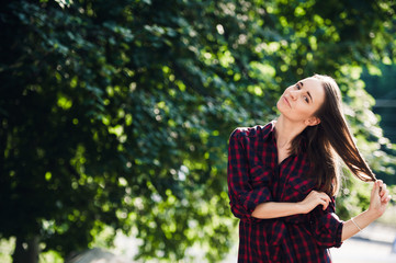 Fototapeta premium Cute teenage girl in casual dress playing with her braids, looking at camera and smiling, standing against green foliage