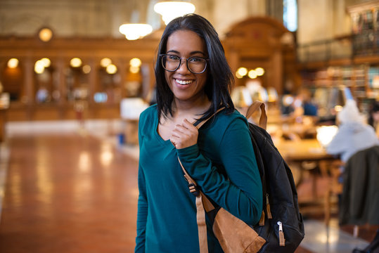 Happy college student girl with backpack in the library smiling. Beautiful young student woman in reading room. Education, smile, study, learning
