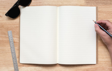 Top view of open notebook with two blank pages, hand hold pencil, sun glasses and ruler on wooden table