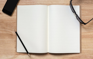 Top view of open notebook with two blank pages, hand hold pencil, sun glasses and smart phone on wooden table