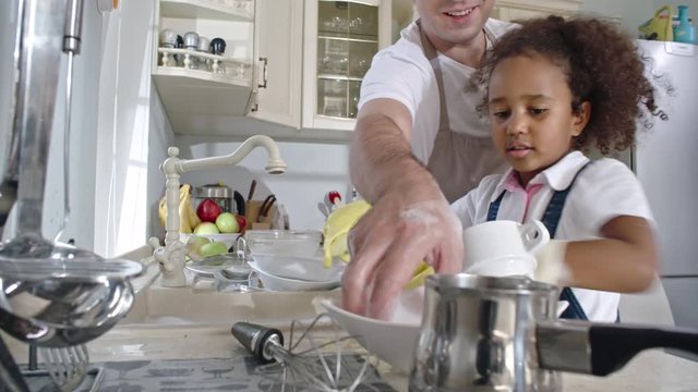 Smiling father passing dishes to little cute daughter standing at sink and washing plates