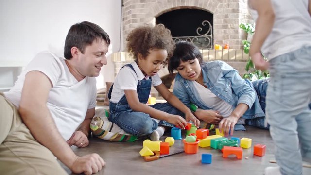 Little African girl sitting on floor in the living room between parents and playing with toy blocks while baby boy walking around them