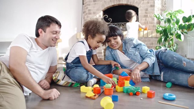 Beautiful mom, dad and little daughter playing with toy blocks while baby boy walking around them in the living room