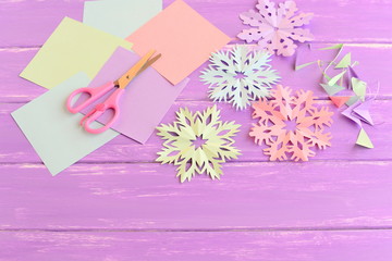 Pink, green, blue and purple paper snowflakes. Paper snowflake set, colored paper sheets and scraps, scissors on wooden background. Kids winter crafts