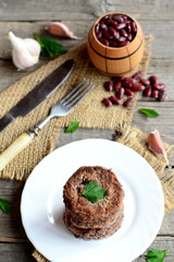 Fried bean burgers with garlic and spices on a plate. Small decorative barrel with uncooked red beans, garlic, parsley, fork, knife on wooden background. Healthy vegetarian food. Rustic style. Closeup