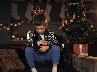 A boy sits on a couch and hugging a cat.