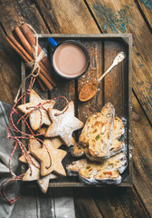 Cocoa in mug with Christmas gingerbread star shaped cookies and pieces of Stollen cake in wooden tray over rustic wooden background, top view, vertical composition