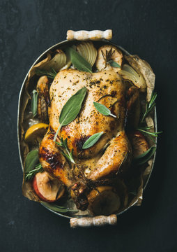 Oven roasted whole chicken with onion, apples and sage in metal serving tray over dark stone background, top view, selective focus. Celebration food concept