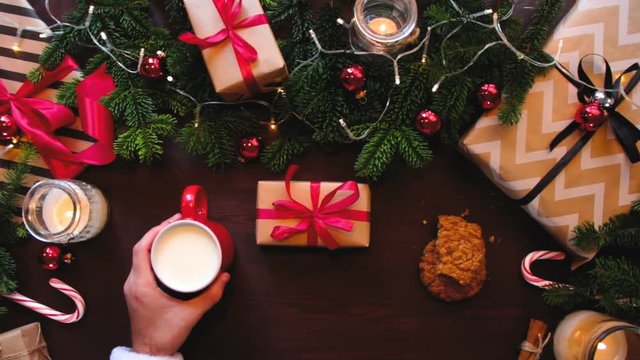 Top hands Santa Claus put a mug with milk and cookies on the table and straightens the ribbon on a gift.