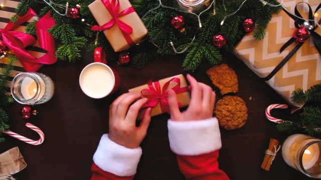 Top hands Santa Claus corrects the red ribbon on the gift and eating cookies with milk.
