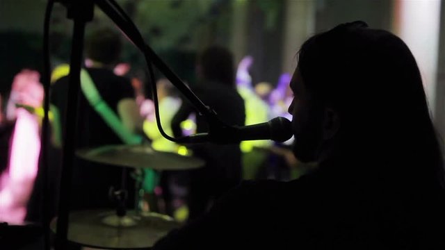 Man singing and playing drums same time live concert. Back side view of silhouette backing vocalist drummer performing solo in microphone with crowd of people jumping happy audience blurry background