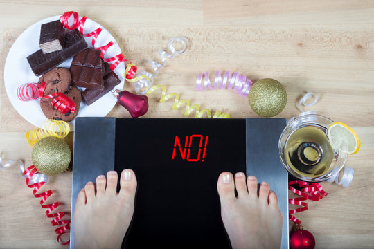 Digital scales with female feet on them and sign"no!" surrounded by christmas decorations, sweets and alchohol. Shows consequences of overeating during Christmas holidays. 