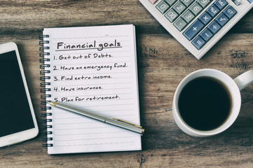 Financial goals on notepad on blank notepad with calculator, coffee, pen and smart phone on a wooden table, retro style. - Powered by Adobe