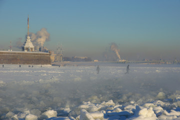 Cold winter day in the city of St. Petersburg. View of Naryshkin Bastion, Peter and Paul Fortress through the steam rising over ice channel to navigate the frozen Neva river. People walk on the ice.  - 129590918