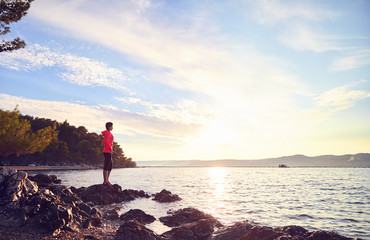 Fototapeta premium Runner resting from routine exercise. Happy jogger relaxing at the beach after run, standing on the stones and looking at the sea. Active young woman enjoying sunrise.