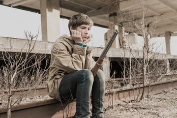 young boy sitting on the railroad tracks near the abandoned train station. wandering boy with a gun. man in a protective cloak with a hood. Post apocalypse. traveling on foot in a post-apocalyptic