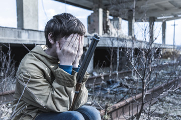 Fototapeta premium young boy sitting on the railroad tracks near the abandoned train station. wandering boy with a gun. man in a protective cloak with a hood. Post apocalypse. traveling on foot in a post-apocalyptic