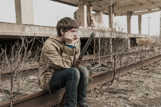 young boy sitting on the railroad tracks near the abandoned train station. wandering boy with a gun. man in a protective cloak with a hood. Post apocalypse. traveling on foot in a post-apocalyptic