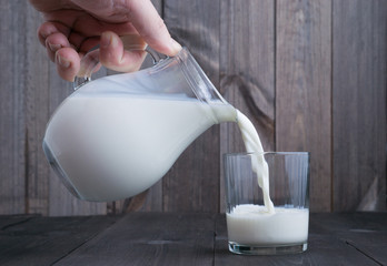 hand woth milk jug pour milk to transparent glass on wooden background