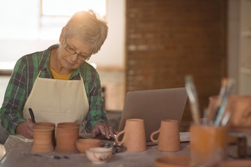 Female potter writing on a book while using laptop