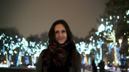 attractive woman at snowy Christmas night smiles looking at the camera in front of park trees decorated sparkling lights