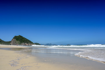 The coast of Atlantic Ocean at Leme beach with Forte Duque de Caxias, mountains, bright blue sky and blurry people on the background at sunny day, Rio de Janeiro, Brazil