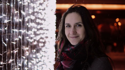 young attractive woman in the falling snow at Christmas night looking at the camera lights at background