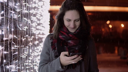 young attractive woman talking on the phone in the falling snow at Christmas night standing near lights wall,