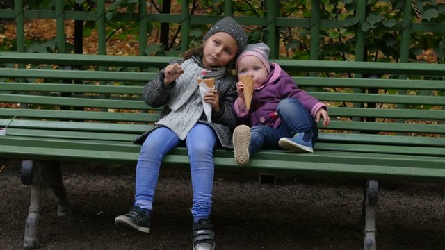 Two sisters sitting on the bench and eating ice-cream. Little girls sit in the park and eating ice cream. Children funning